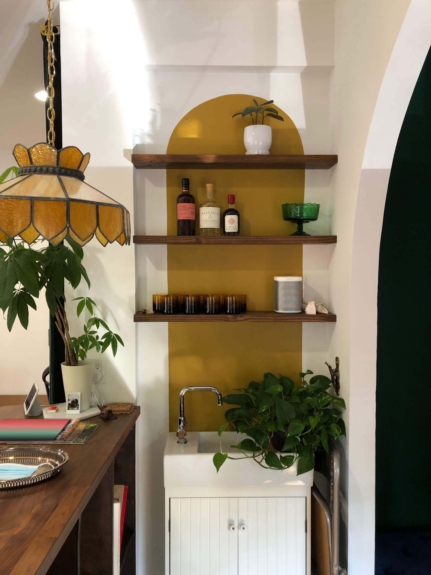Floating shelves with plants, cocktail glasses, and whiskey bottles, with a painted gold archway behind them